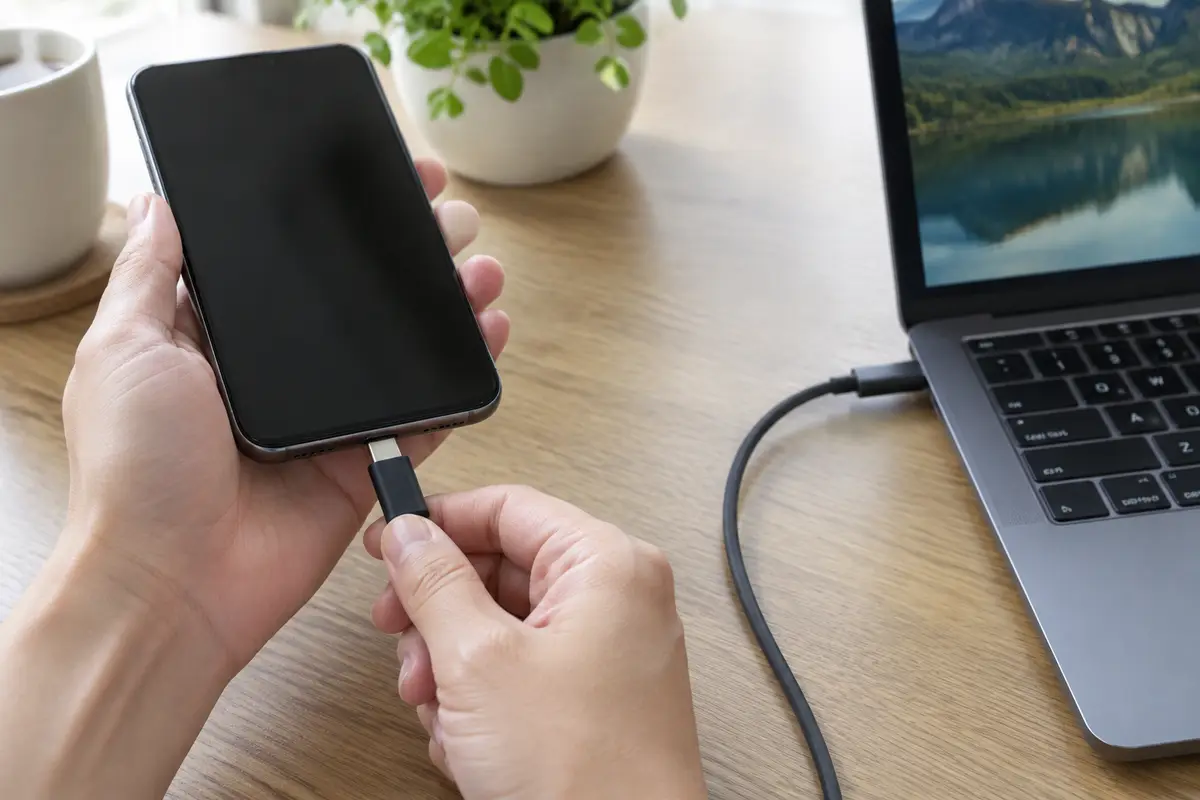 Руки вставляют штекер USB-C в нижнюю часть смартфона, второй конец кабеля идёт к ноутбуку на столе